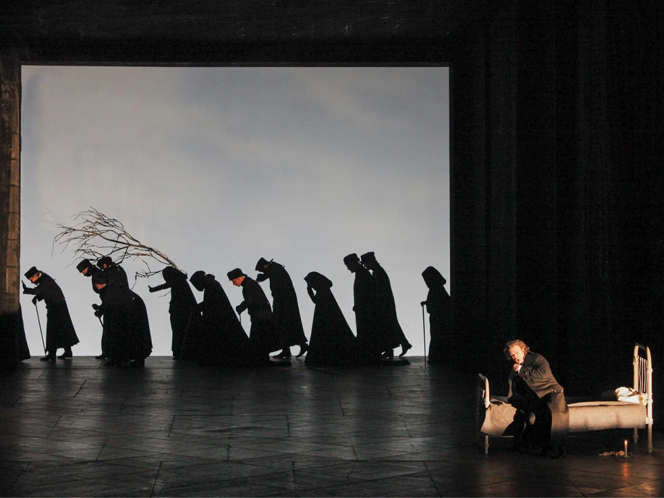 A group of people wearing dark clothing and hats walk while hunched over, holding branches, on a stage. One person is seated on a bed in the foreground, facing the audience.