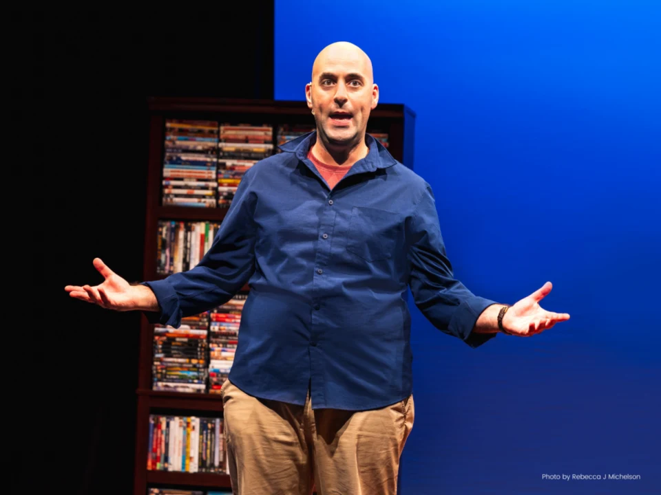 Production photo of Clowns Like Me in New York, featuring Scott Ehrenpreis wearing a blue shirt and beige pants, standing with his arms outstretched on a stage with a bookshelf in the background.