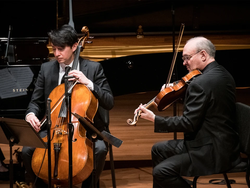 Two musicians in formal attire perform on stage. One plays the cello while the other plays the viola, with a grand piano visible in the background.