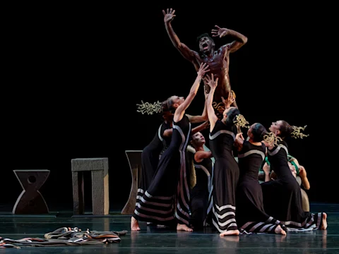 A group of dancers in black costumes reaches upward toward a male dancer elevated above them on stage, with abstract set pieces in the background.