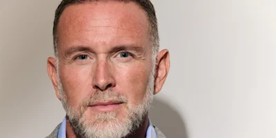 Close-up of a middle-aged man with short hair and a gray beard, wearing a light blue shirt and gray blazer, looking directly at the camera against a plain light background.