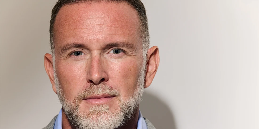 Close-up of a middle-aged man with short hair and a gray beard, wearing a light blue shirt and gray blazer, looking directly at the camera against a plain light background.