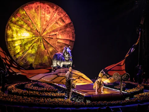 Colorful stage with performers, a large wheel backdrop, and a realistic animal figure, surrounded by an audience and yellow floral decorations.