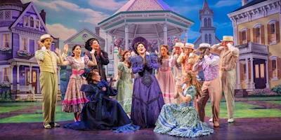 A theatrical cast in colorful period costumes performs on stage in front of a backdrop depicting Victorian-style houses and a gazebo.