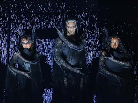 Three people in elaborate black costumes and dramatic makeup pose with arms crossed against a backdrop of falling white digital lines.