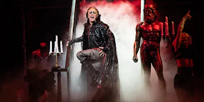 Performer in dramatic costume stands center stage, flanked by a shirtless man holding candles and a woman, with heavy fog and theatrical lighting.