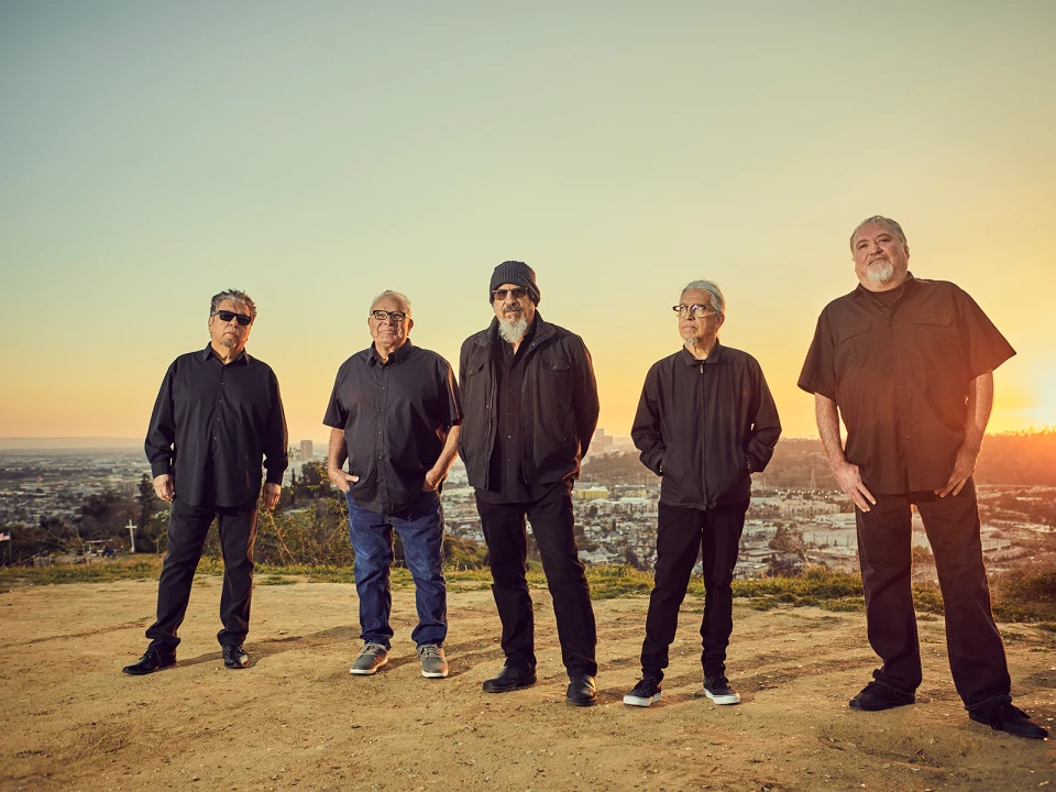 Five men stand outdoors at sunset, with a cityscape in the background. They are casually dressed, and most have their hands in their pockets.