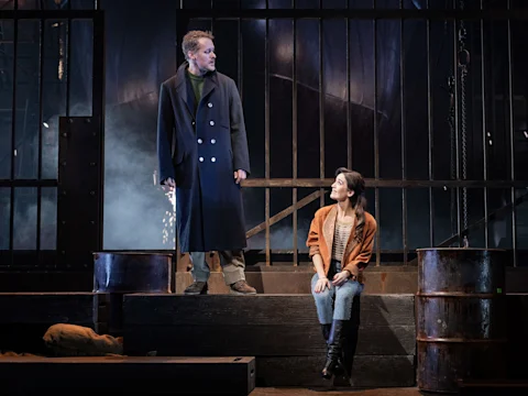 A man in a dark coat stands on a raised platform, looking at a seated woman in casual clothes; they are on a dimly lit stage with metal bars and barrels in the background.
