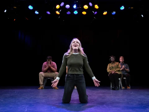 Production shot of Washington Improv Theater: Genre Reveal Party in Washington, showing a woman kneels under colorful lighting, with three seated individuals in the background.