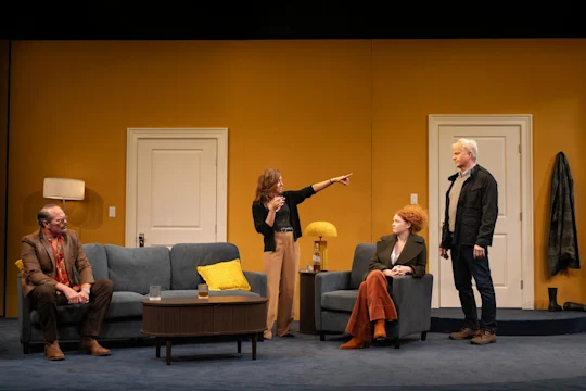 Production photo of Let's Love! in New York, showing four people are in a living room with yellow walls; one woman stands and points while the others, seated or standing, look at her. The scene appears to be from a staged play.