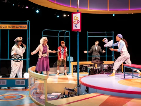 A colorful stage scene featuring five actors in various costumes, including a captain, an elderly woman, a young man, a person in formal attire, and someone in casual attire, amidst vibrant set pieces.