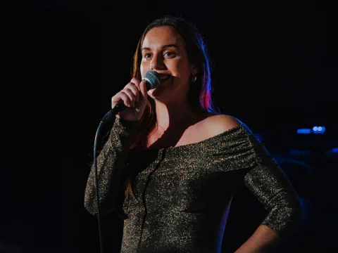 A woman in a sparkly off-the-shoulder top stands on stage, holding a microphone and speaking or singing.