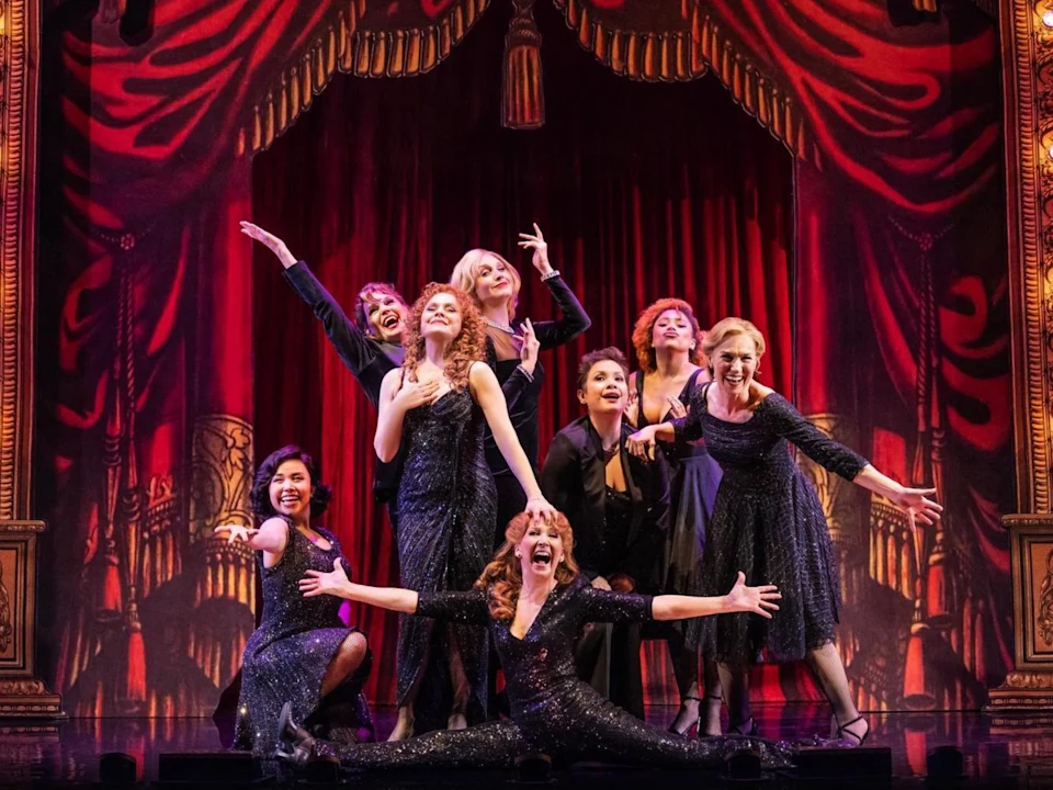 A group of performers in dark, sparkly outfits strikes various poses on stage with a red, ornate curtain backdrop. Bernadette Peters Lea Salonga and the company of STEPHEN SONDHEIM'S OLD FRIENDS at the Ahmanson Theatre. 