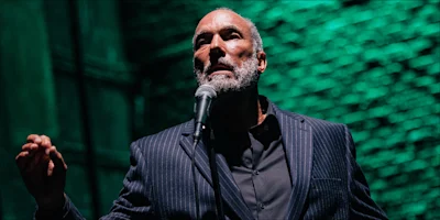 A man in a pinstripe suit stands in front of a microphone with a green-lit brick wall behind him, gesturing with one hand.