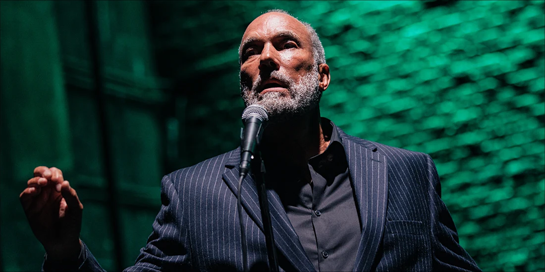 A man in a pinstripe suit stands in front of a microphone with a green-lit brick wall behind him, gesturing with one hand.