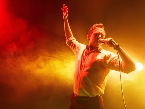 A man sings into a microphone on stage, raising one arm, with dramatic red and yellow lighting and smoke in the background.