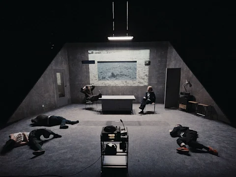 A dimly lit room with a desk in the center, two people seated, and three people lying on the floor. A projector displays a grayscale image on the back wall.