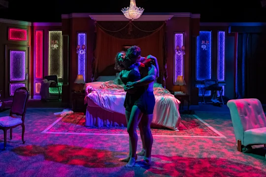 Two people embrace in the center of a dimly lit, ornate bedroom with colorful lighting and an unmade bed in the background.