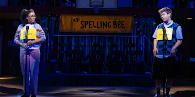 Two contestants stand at microphones onstage during a spelling bee, with a yellow "Spelling Bee" banner hanging in the background.