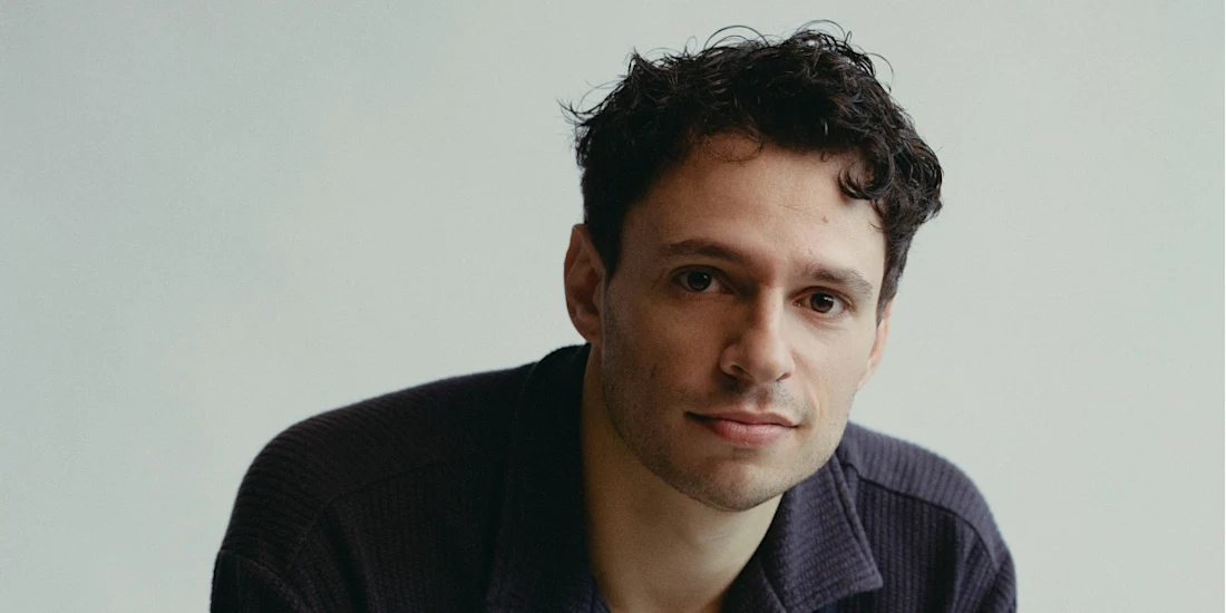 A man with short, curly dark hair and light skin looks at the camera, wearing a dark collared shirt against a plain light background.