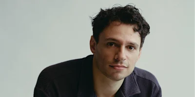 A man with short, curly dark hair and light skin looks at the camera, wearing a dark collared shirt against a plain light background.