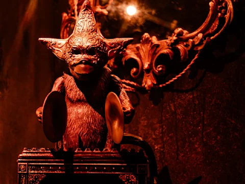 A mechanical monkey statue with cymbals and an ornate mask sits in front of a decorative mirror, illuminated by warm, reddish lighting.