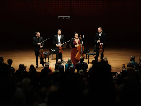 Four musicians holding string instruments stand on stage facing an audience, appearing to acknowledge applause after a performance.