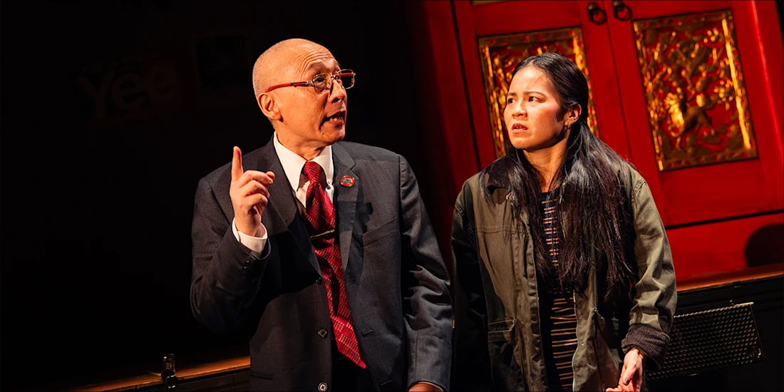 An older man in a suit points upward while speaking to a younger woman wearing a jacket in front of a red decorative door.