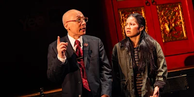 An older man in a suit points upward while speaking to a younger woman wearing a jacket in front of a red decorative door.