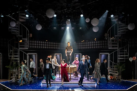 A group of actors in formal attire perform on stage, with one person elevated on a platform under a spotlight. The set features spiral stairs, balloons, and dramatic lighting.
