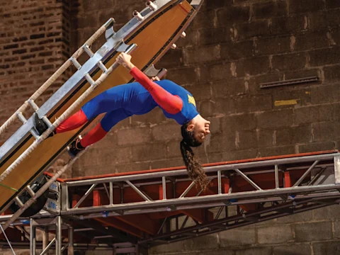 An acrobat in a blue and red leotard hangs upside down while gripping an angled, wooden obstacle on a gym course.