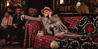 A man in a plaid jacket and hat lounges on a patterned sofa, talking on a rotary phone, while several people seated in the background watch him.