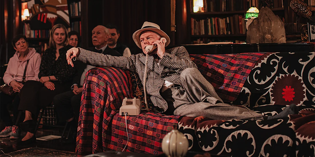 A man in a plaid jacket and hat lounges on a patterned sofa, talking on a rotary phone, while several people seated in the background watch him.