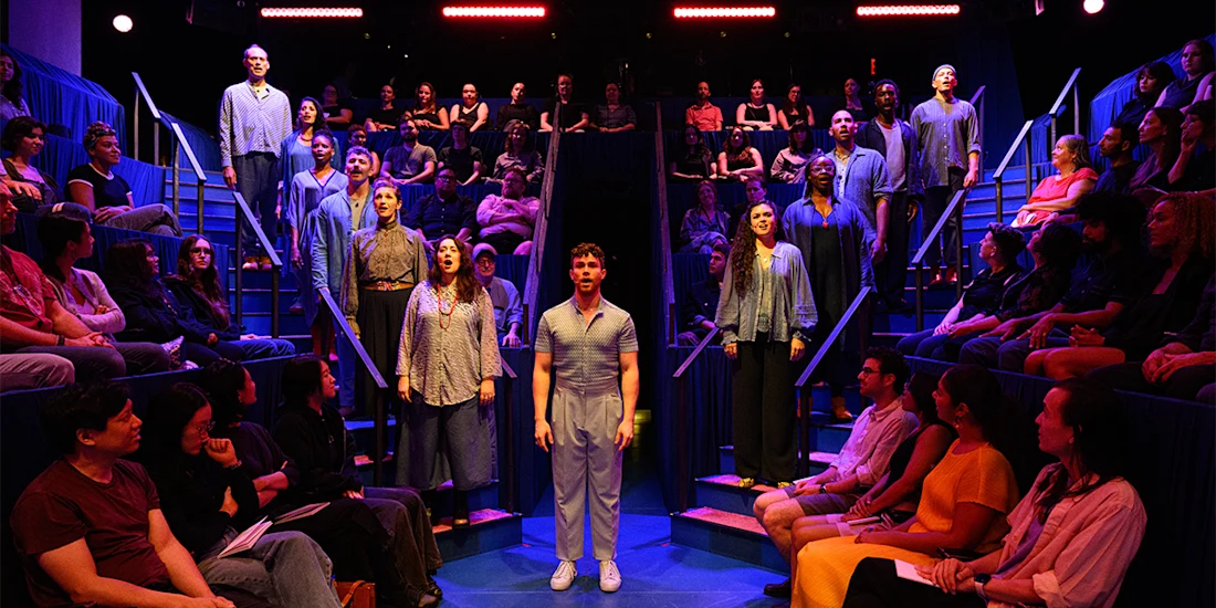 A group of performers stand on stage surrounded by an audience seated on tiered blue benches, under bright stage lights.