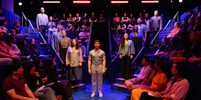 A group of performers stand on stage surrounded by an audience seated on tiered blue benches, under bright stage lights.