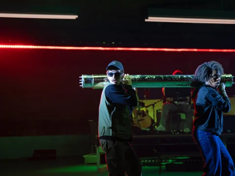 Two performers on stage; one holds a large metallic tube over their shoulder, wearing sunglasses and a cap. A band is visible in the background with red and green lighting.