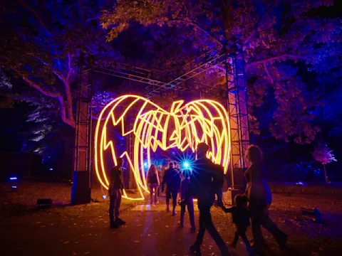 People walk under a large illuminated pumpkin-shaped archway at night, surrounded by trees and vibrant blue and orange lighting.