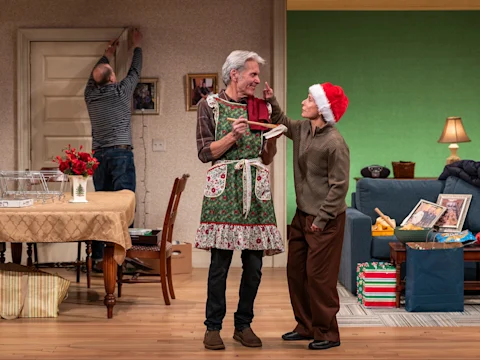 Three people in a living room decorated for Christmas; one is adjusting a photo on the wall, another in an apron holds a spoon, and a third in a Santa hat touches the apron wearer's nose.