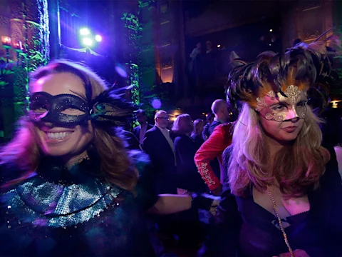 Two women in elaborate feathered masks stand in a dimly lit, crowded ballroom with colorful lights and ornate decor in the background.