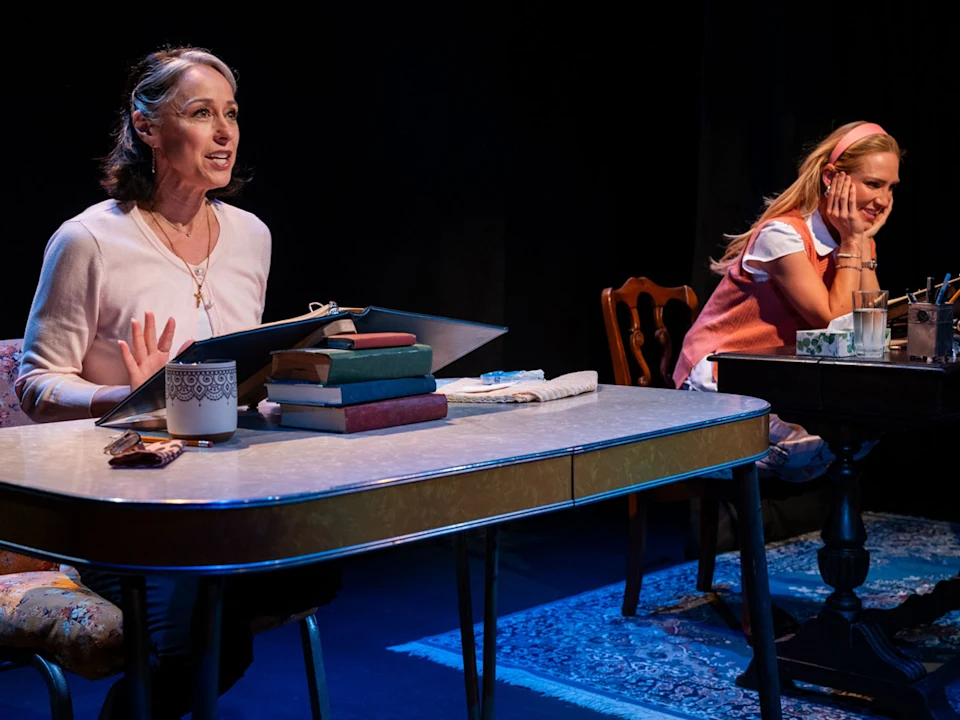 Two women sit at separate desks onstage, each with books and papers, engaged in a dramatic performance under stage lighting.