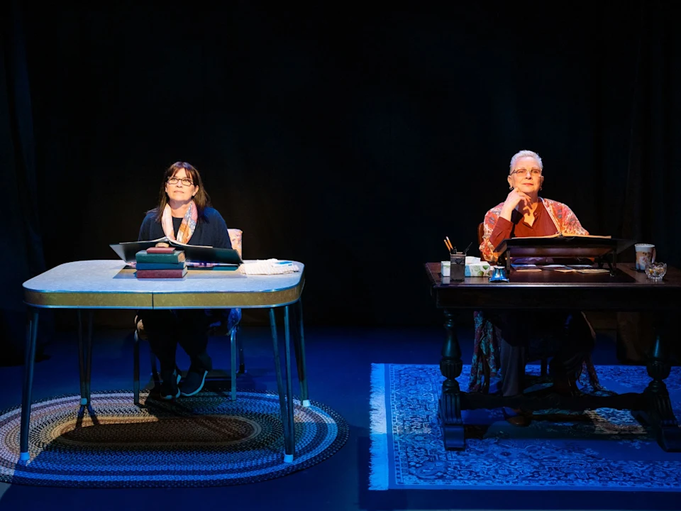 Two women sit at separate desks on a stage, each with papers and writing materials, under individual spotlights against a dark background.