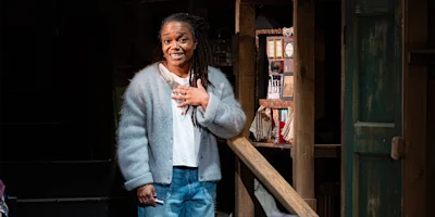 A woman with long braids, wearing a light gray cardigan and jeans, stands indoors by a wooden railing, resting one hand on her chest and smiling.