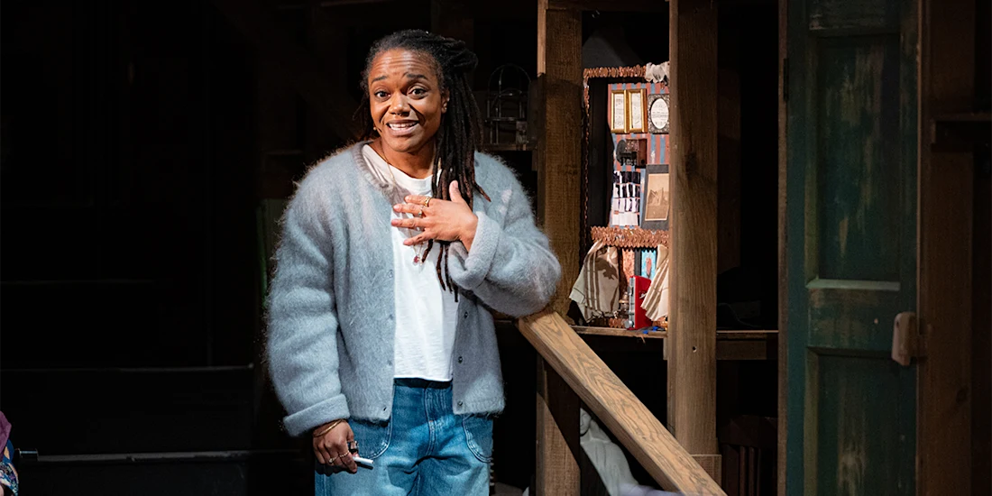 A woman with long braids, wearing a light gray cardigan and jeans, stands indoors by a wooden railing, resting one hand on her chest and smiling.