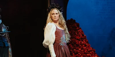 A woman in a long dress with white sleeves and a black floral crown stands on stage, with a backdrop of red roses and a blue circular shape behind her.
