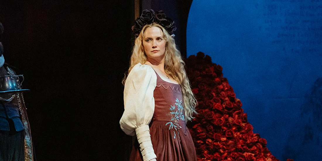 A woman in a long dress with white sleeves and a black floral crown stands on stage, with a backdrop of red roses and a blue circular shape behind her.
