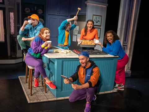 Six people in colorful outfits gather around a kitchen island; one holds a mallet, another presents a baked dish, while others display cheerful, animated expressions.