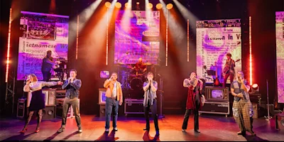 Seven performers sing and play instruments on a brightly lit stage with projected newspaper headlines and a band in the background.