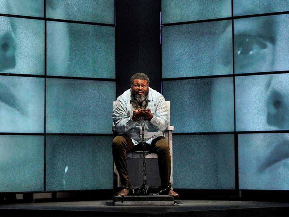 A man sits on a chair with chains around his wrists and ankles. Multiple screens in the background display a solemn face.