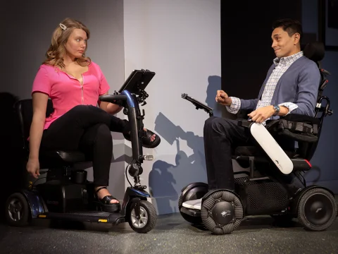 A woman and a man, each using a modern mobility scooter, are engaged in a conversation indoors.