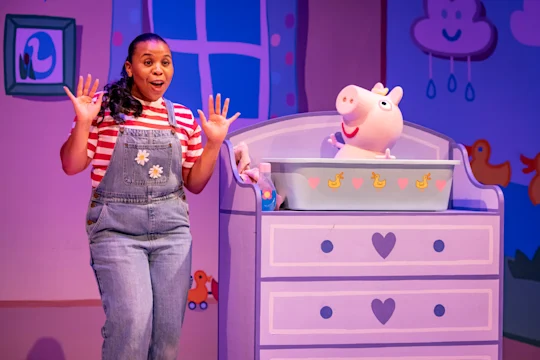 A woman in overalls stands with raised hands beside a crib with an Evie puppet, set in a colorful, cartoon-themed room.
